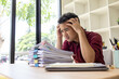 © Kield KD - A man in a red shirt is sitting at a desk with a pile of papers in front of him. He is stressed and overwhelmed by the amount of work he has to do