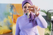 © Studio Marmellata - A happy young woman wearing a purple hijab presents a set of keys to the camera. She stands in her bright art studio, celebrating a new beginning.