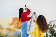 © Studio Marmellata - Two women work together on home improvement. One woman stands on a ladder using a power drill to make a hole in the wall while the other assists her. They are preparing to hang artwork.