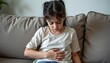 © K-MookPan - Sad child sitting on couch with hand on stomach showing signs of discomfort and distress at home indoors