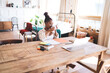 © BullRun - Woman working remotely from stylish home interior, reaching toward laptop with one hand while sitting at rustic desk filled with planners, markers, phone and digital accessories