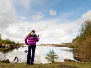 Wall Mural - Girl tourist enjoys view on a lake with cloudy sky reflection. Travel and tourism concept. Water is calm and green bushes and trees growing on the lake bank. Calm relaxed mood. Voyage and adventure.