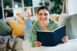 © deagreez - Happy young woman reading a book at home, enjoying a cozy and relaxing day in a comfortable living room during daylight.