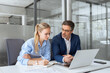 © Stock 4 You - Two partners, team of professional business people working, studying, writing notes sit at desk. Mature focused senior mentor leader man showing young woman manager project on laptop in modern office