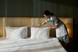 © pressmaster - Hotel housekeeper making bed in a clean, modern room, arranging pillows on neatly made bed, wearing uniform, and focused on task in a well-lit environment