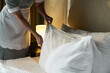 © pressmaster - Housekeeper arranging white bedding in a well-lit hotel room while straightening pillows and ensuring neatness and cleanliness for guests' comfort