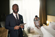 © pressmaster - Portrait of hotel manager in suit holding tablet while housekeeping staff making bed in background. Manager is standing in modern, comfortable guest room