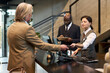 © pressmaster - Businessman in beige suit interacting with hotel reception staff while checking in at front desk. Receptionist scanning card while another staff member taking notes on clipboard