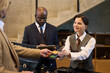 © pressmaster - Concierge processing a payment at hotel front desk with two clients present. Person in background holding tablet and dressed in business attire