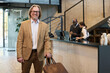 © pressmaster - Smiling businessman holding bag while standing in hotel lobby with front desk staff attending customers in background