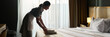© pressmaster - Housekeeper arranging neatly folded towels on a bed during cleaning routine in a hotel room with white curtains and warm lighting