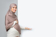 © Prostock-studio - Pregnant muslim woman in hijab pointing at white advertisement board with free space, touching her belly and looking at camera, grey background