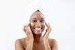 © Prostock-studio - Skin hydration, anti-aging care and home spa. Cheerful attractive millennial african american woman with towel on head after shower applies cream on face, in bedroom in morning, on white background