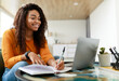 © Prostock-studio - Note Taking Concept. Smiling young African American woman sitting at desk working on laptop and writing letter in paper notebook, holding pen and looking at screen. Happy millennial female using pc