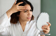 © Prostock-studio - Upset arabic young woman in white silky robe looking at hand mirror, checking her skin, touching her forehead with red pimple, home interior, closeup photo. Acne, pimples, dull skin