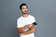 © SHOTPRIME STUDIO - Handsome young man with smartphone looks thoughtfully at the camera, wearing a casual white t-shirt against a solid grey background. Lifestyle concept.