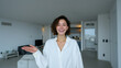 © Favio - Young woman welcoming viewers inside bright modern apartment interior with white walls and cozy living space suitable for real estate rental or home sales promotion.