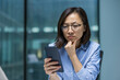© Liubomir - A thoughtful Asian woman wearing glasses examines her smartphone, pondering a message in a modern office setting.