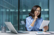 © Liubomir - An asian businesswoman is thoughtfully reviewing information on a tablet while sitting at her desk in a modern office setting. There is a laptop.