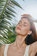 © SHOTPRIME STUDIO - woman enjoying sunlight with closed eyes near tropical leaves, natural beauty, peaceful mood, summer vibes, clear skin glowing softly under bright sun