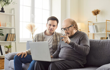 Young man, adult son teaching interested mature senior father using laptop computer online applications at home, watching photo video content in social network, shopping in internet store, modern tech