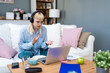 © Srdjan - Young woman wearing headphones having video call on laptop, sitting on cozy couch at home, working remotely or studying online, gesturing while talking, surrounded by coffee, notes, and documents.