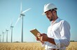 © Pete - Engineer wearing white hard hat, shirt, checks wind turbine production using tablet device. Man stands in golden wheat field, in front of wind turbines. Eco friendly energy, sustainable clean power.