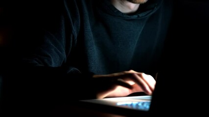 Anonymous person in a hoodie is typing on a laptop in a dark room with blue screen light illuminating their face and keyboard