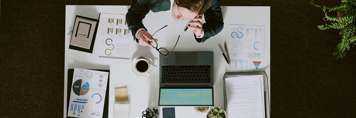 Wall Mural - Business person analyzing documents and reports while sitting at desk filled with papers, charts, and laptop in office setting