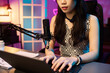 © pressmaster - Asian woman working on laptop in dimly lit modern office with professional microphone and stylish decor, focused on recording vlog without looking at camera