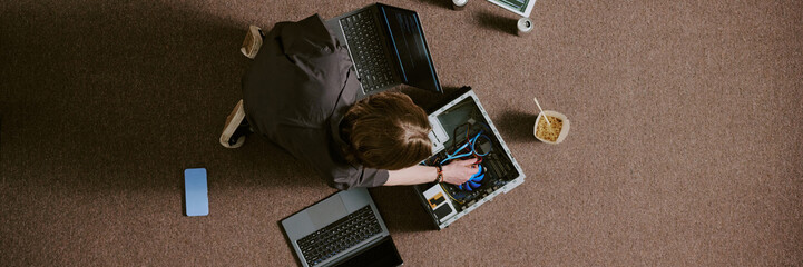 Wall Mural - Technician repairing multiple laptops while sitting on carpeted floor, using various tools and devices, showing in depth focus and technical skillset during task