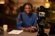 © pressmaster - Host recording podcast episode, seated at desk with microphone and camera capturing. Office elements like coffee cup, notes visible on desk. Hosting engaging discussion
