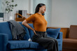 © Parichat - Young woman experiencing back pain while sitting on a sofa in her living room, a common health issue
