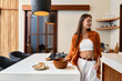© SHOTPRIME STUDIO - Young woman in an orange shirt and white pants, smiling while preparing healthy food in a stylish kitchen, emphasizing a cozy lifestyle and seasonal freshness