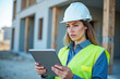 © Sync - Woman construction foreman engineer using digital tablet on job planning site with modern building in background, workplace technology and safety concept with daylight and reflective vest