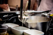 © AokiSuko Studio - Close-up of steaming seafood chowder ladle over bowls in a busy professional kitchen, capturing the dynamic cooking environment. Perfect for culinary, restaurant, and food service marketing.