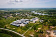 © Raivo - Aerial view of a rural industrial zone near a small town and lake, with warehouse buildings, roads, fields, and surrounding residential areas..