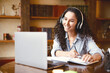 © Prostock-studio - Home Office. Portrait of smiling young lady in wireless headphones sitting at desk, writing in notebook and using computer. Woman watching online tutorial, webinar or seminar, studying at home