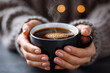 © Nadezda Ledyaeva - Warm cup of coffee held in hands, steam rising from the surface, cozy atmosphere with blurred lights in the background, inviting comfort and relaxation in a serene setting