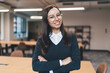 © BullRun - Confident young businesswoman with long dark hair and glasses smiling with arms crossed in open coworking space, expressing leadership, self-assurance and career growth