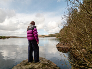 Fototapete - Girl tourist enjoys view on a lake with cloudy sky reflection. Travel and tourism concept. Water is calm and green bushes and trees growing on the lake bank. Calm relaxed mood. Voyage and adventure.