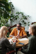 © pressmaster - Group of three people sitting at round table, discussing plans in cozy indoor garden setting, surrounded by lush green plants, look relaxed and engaged in conversation