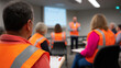 © Curioso.Photography - Group of workers wearing orange safety vests attending a training session or safety presentation in an industrial setting.