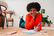 © Jose Calsina - A cheerful young woman is lying on the floor and writing in her notebook, reflecting her creativity and enthusiasm for learning as she engages in productive writing and thought exploration at home.