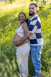 © Bogdan Pictures - A loving pregnant couple enjoys a sunny summer day outdoors, with the husband tenderly hugging his wifes baby bump in a serene setting filled with lush greenery and vibrant flowers