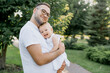 © Bohdan - stylish bearded father with his little daughter in his arms against the backdrop of green trees on a walk in the park hugging a baby