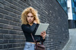 © Srdjan - Young woman with curly blonde hair using digital tablet against modern black brick wall background, standing outdoors, focused and stylish, casual urban lifestyle and technology usage concept.