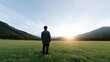 © kiimoshi - A solitary figure stands in a vast green field while gazing at a serene sunrise over distant mountains, embodying tranquility and reflection in nature's beauty.