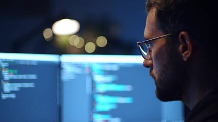Focused male programmer working on multiple computer screens displaying code in a dark office.
