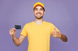 © ViDi Studio - Young delivery guy employee man wear yellow cap t-shirt uniform workwear work dealer courier hold in hand mock up of credit bank card show thumb up isolated on plain purple background Service concept